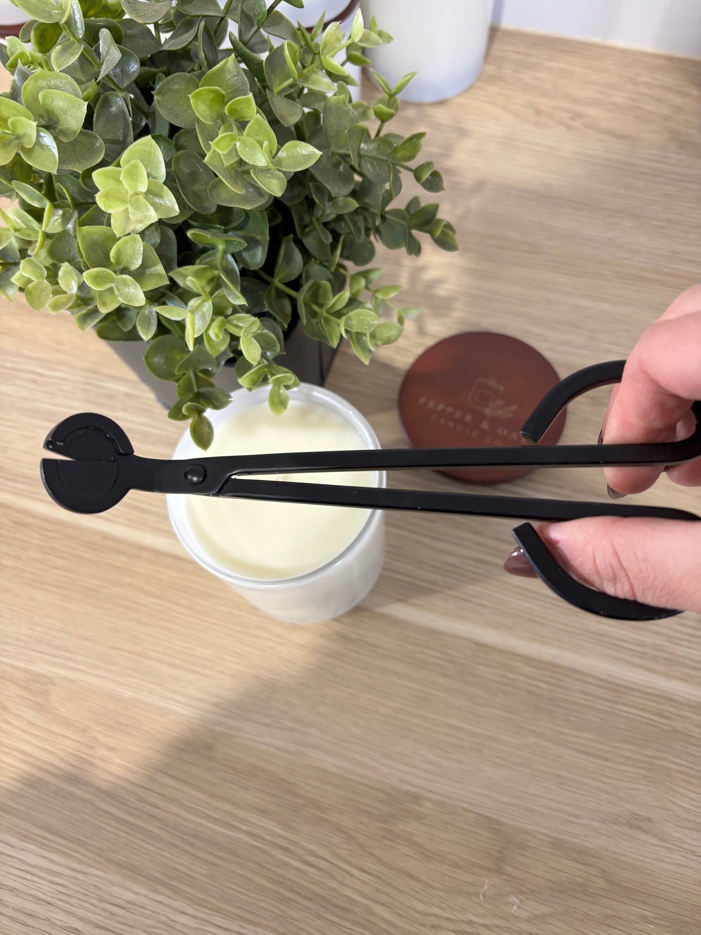 Black wick trimming scissors held over a plant and container on a wooden surface.
