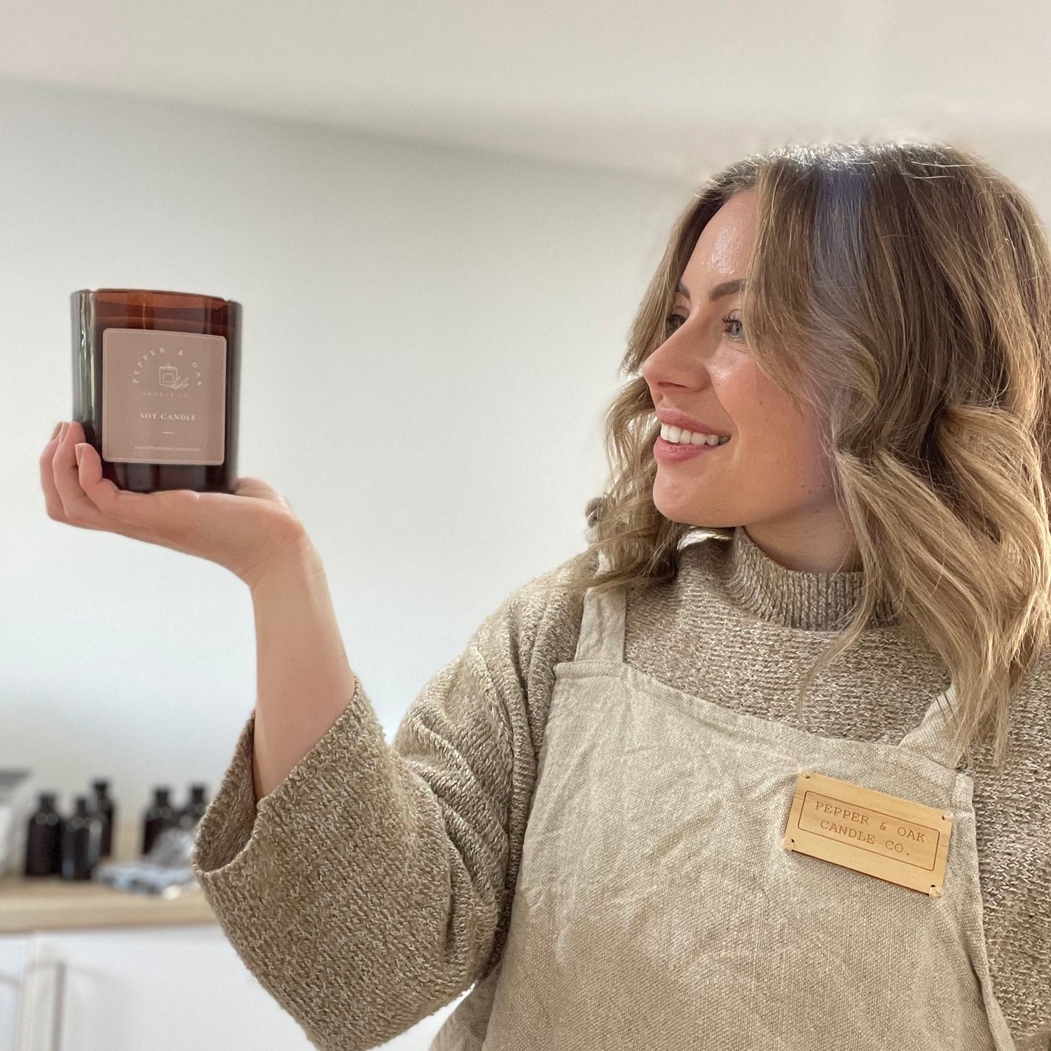 Woman holding a soy candle in a kitchen wearing a branded apron