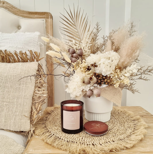 A large soy candle with a vanilla fragrance, placed on a round rattan mat next to a candle holder with a bouquet of dried flowers in a white jar.