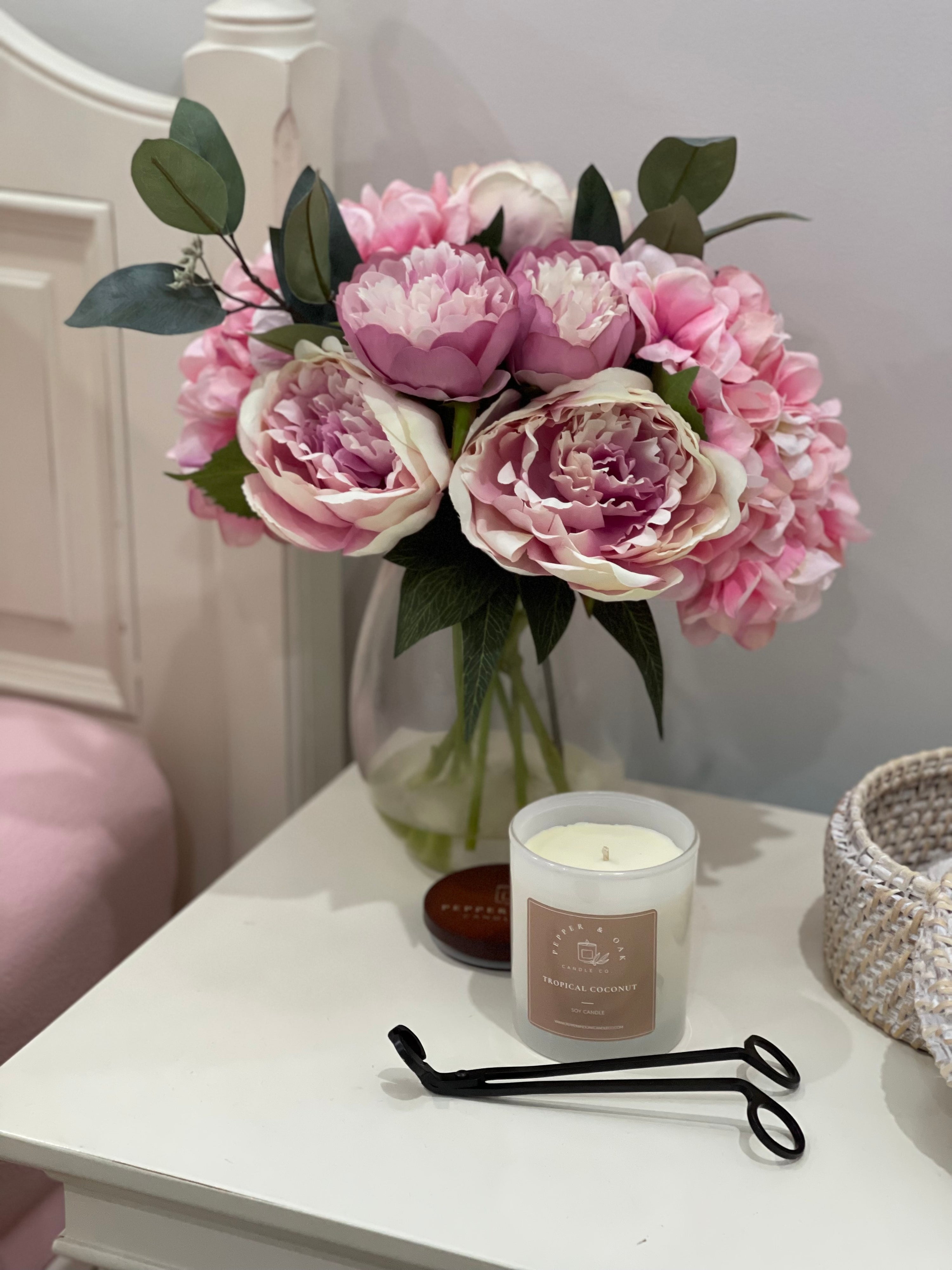 A large tropical coconut fragranced candle on a bedside table beside wick trimmer and pink and purple peonies in a vase